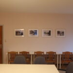 Besprechungsraum im Haus der Begegnung: das Foto zeigt im Vordergrund einen großen weißen Tisch mit gepolsterten Stühlen, dahinter eine weitere Wand zusehen mit Schreibtischen und Stühlen, darüber gerahmte Siebdrucke mit Dorfmotiven, rechts ein Fenster mit Rollo, links eine Holztür und ein Flipchart