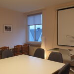 Besprechungsraum im Haus der Begegnung: das Foto zeigt im Vordergrund einen großen weißen Tisch mit gepolsterten Stühlen, dahinter ein Schreibtisch mit Stuhl und Lampe an der Wand, darüber eine große Leinwand, links daneben eine große Leinwand für Projektionen, links daneben eine Stehlampe und ein Fenster mit Rollo, links ist eine weitere Wand zusehen mit Schreibtischen und Stühlen, darüber gerahmte Siebdrucke mit Dorfmotiven